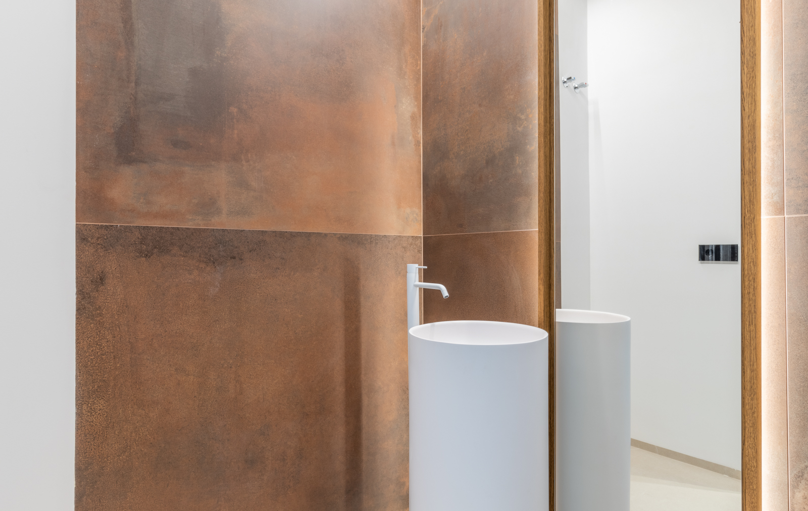 Detalle de baño con lavabo cilíndrico blanco y pared de azulejos color óxido con espejo retroiluminado.