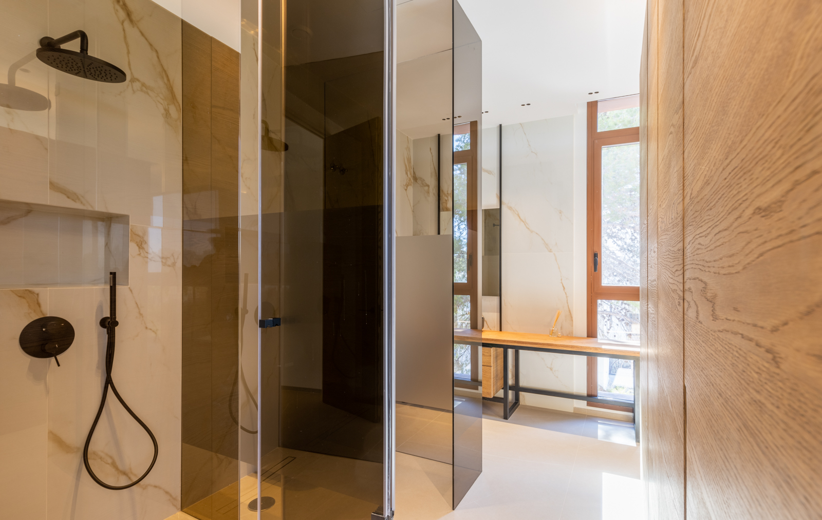 Detalle de ducha con mampara ahumada y tocador de madera clara en baño moderno con azulejos veteados.