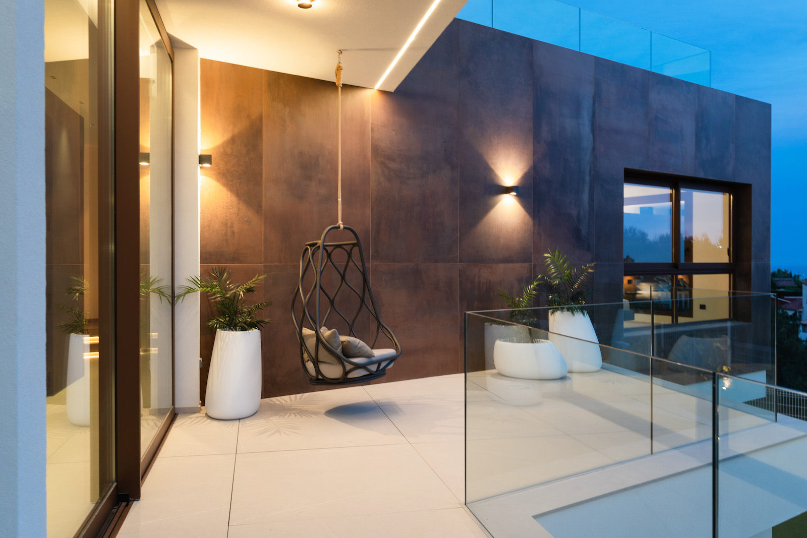 Terraza con pared de azulejos color óxido, silla colgante y barandilla de vidrio.
