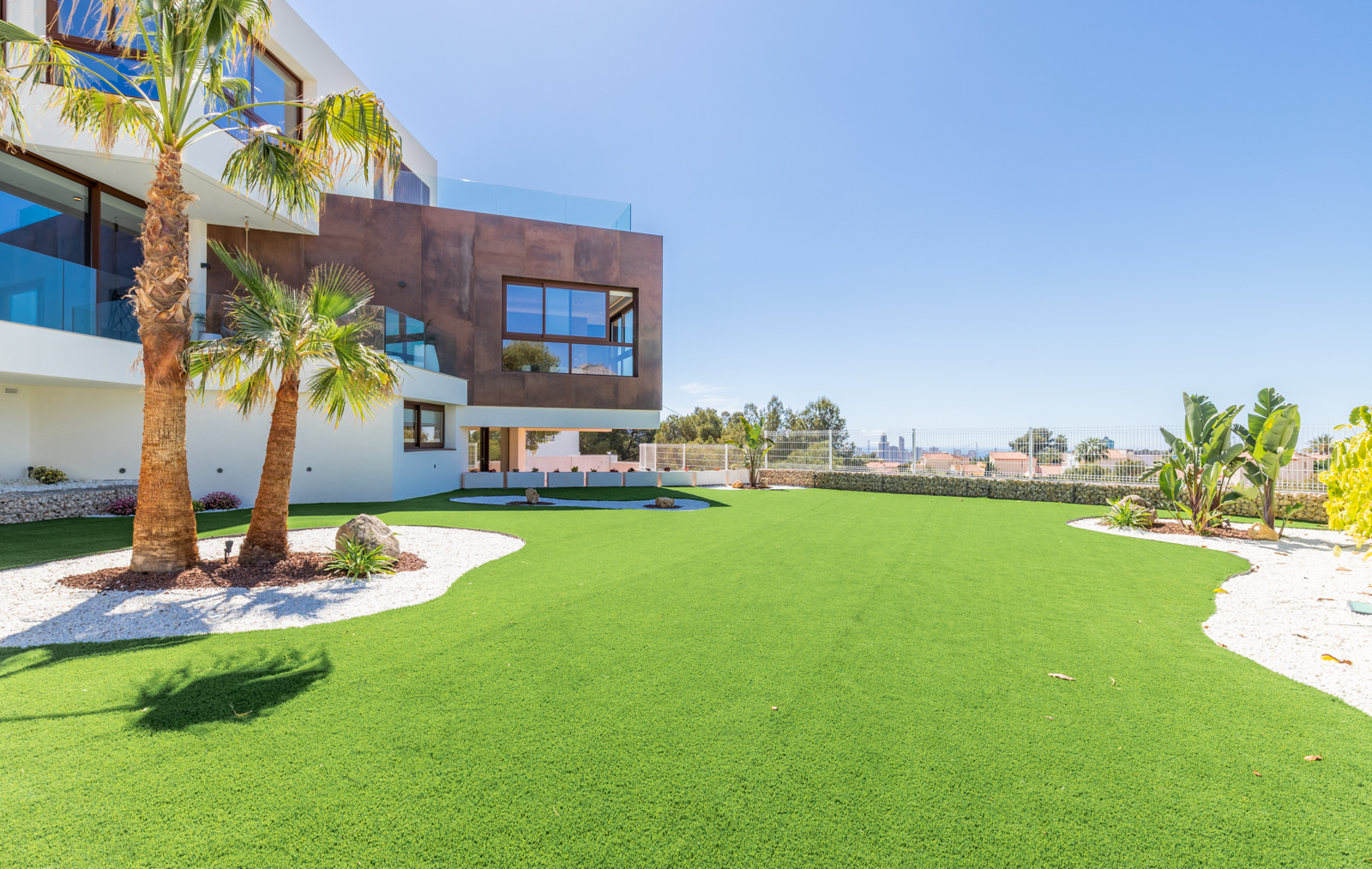 Vista del jardín con césped artificial, palmeras y fachada moderna en La Nucía.