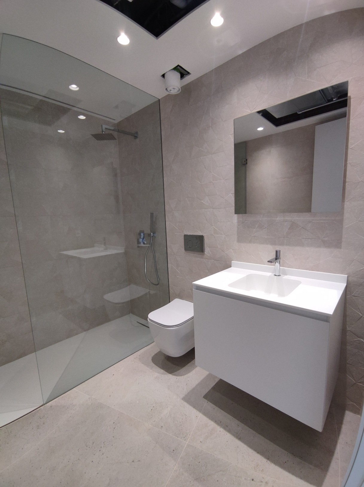 Baño moderno con ducha de obra acristalada, inodoro suspendido y mueble de lavabo flotante blanco sobre revestimiento cerámico con relieve geométrico.