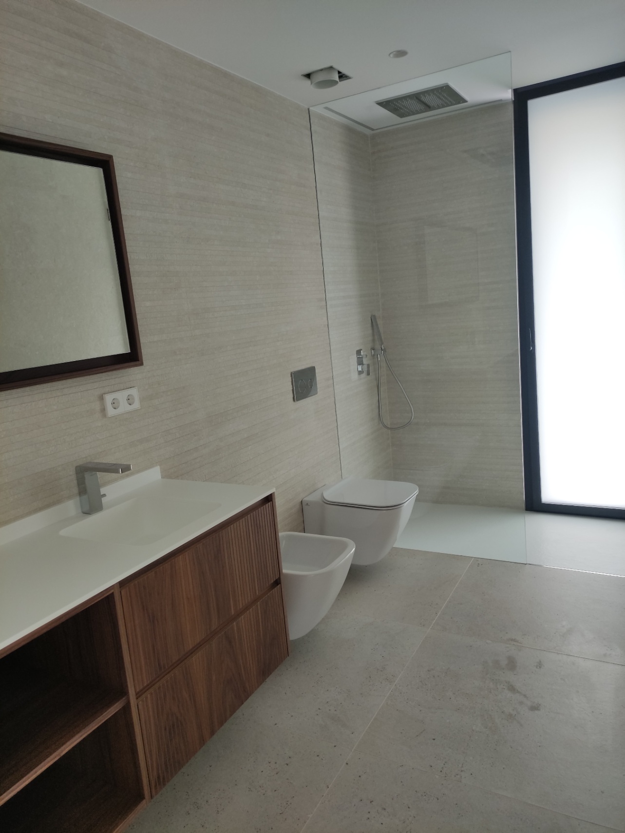 Cuarto de baño con mueble de madera natural, lavabo integrado, sanitarios suspendidos y zona de ducha con rociador empotrado en el techo y mampara de cristal.