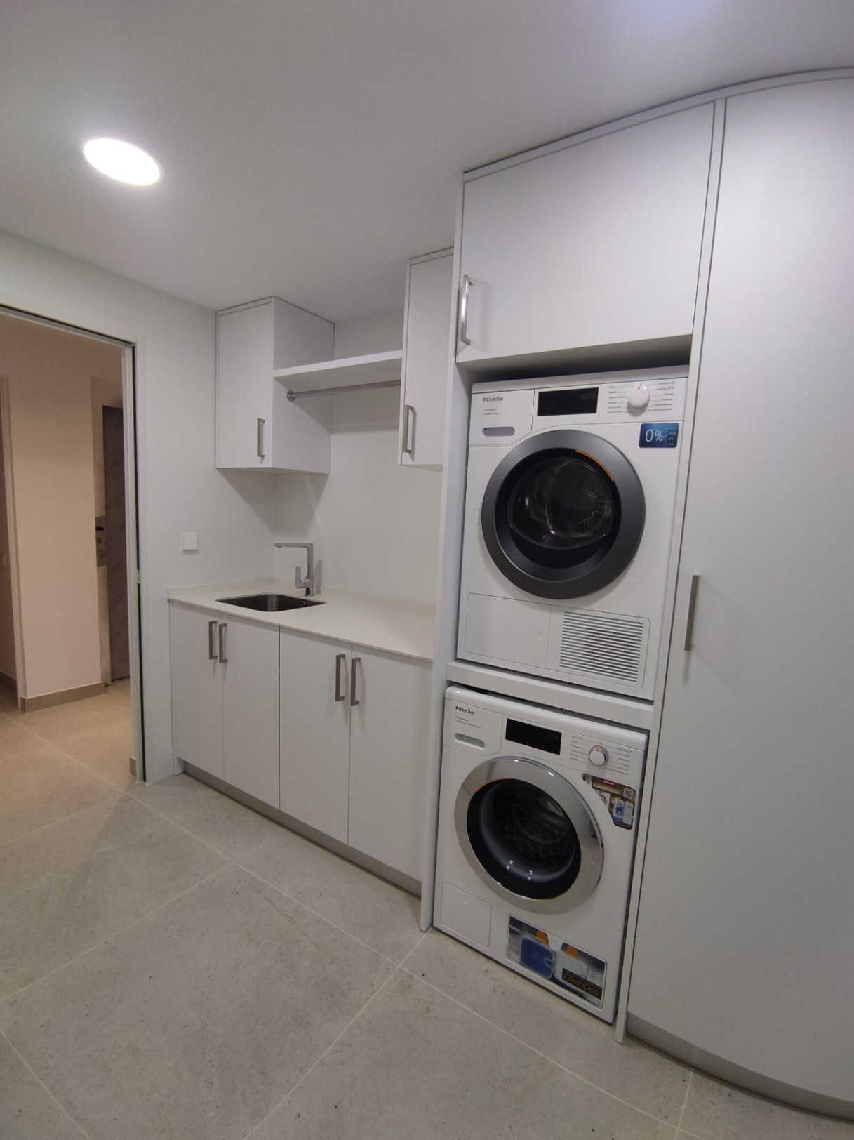 Cuarto de lavado funcional con electrodomésticos Miele en columna, armarios blancos minimalistas, fregadero integrado y suelos de gres porcelánico en tonos neutros.