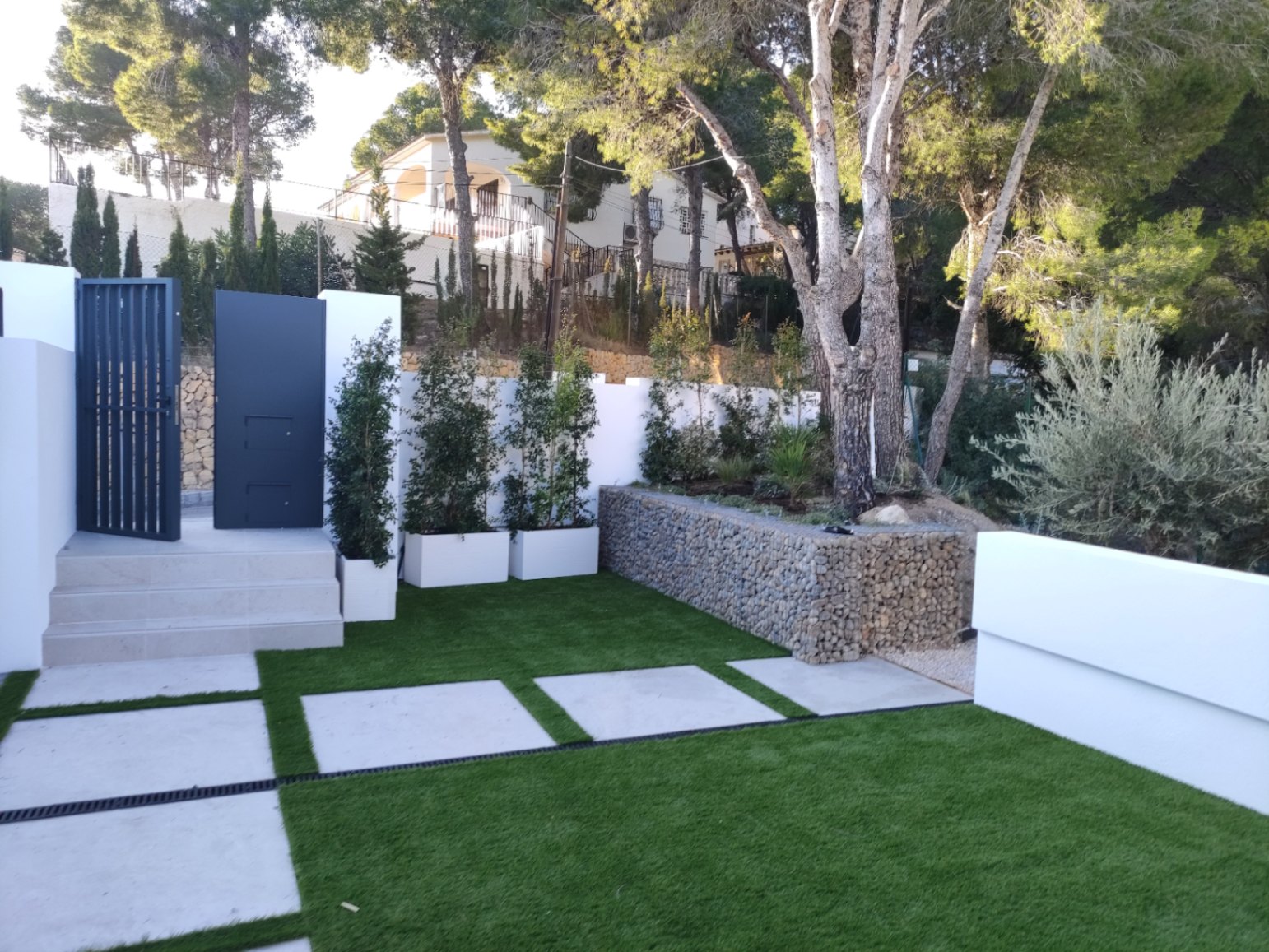 Entrada exterior con portón metálico moderno, peldaños de piedra clara y jardín paisajístico con césped artificial, losas de hormigón y un muro de gaviones decorativo.