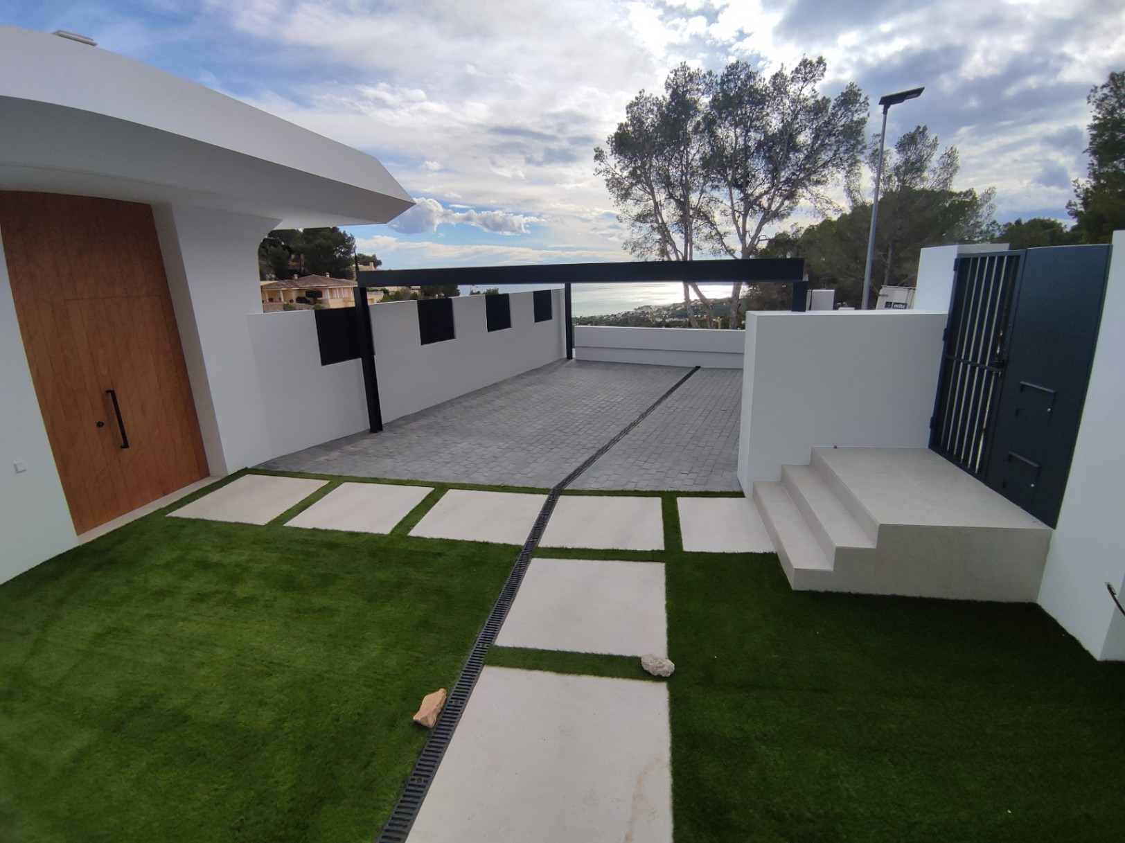 Entrada exterior con puerta de madera noble, zona de aparcamiento pavimentada y jardín de césped artificial con losas blancas. La arquitectura moderna ofrece vistas al mar.