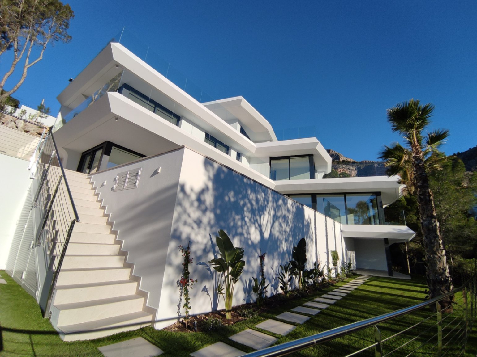 Fachada de diseño contemporáneo distribuida en varios niveles con terrazas de cristal, escalera exterior y jardín con camino de piedra y palmeras.