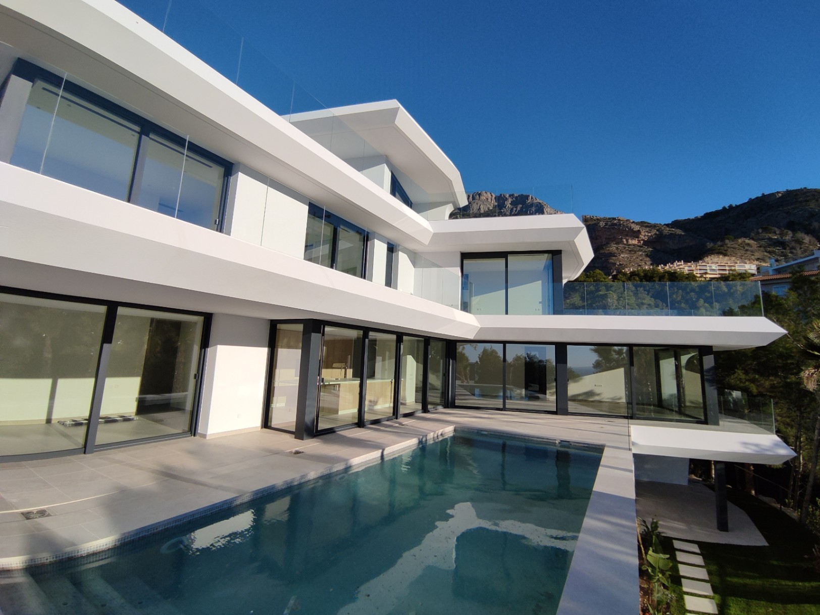 Fachada moderna de líneas blancas con amplios ventanales, terrazas con barandillas de cristal y una piscina infinita integrada frente a las montañas de Altea Hills.