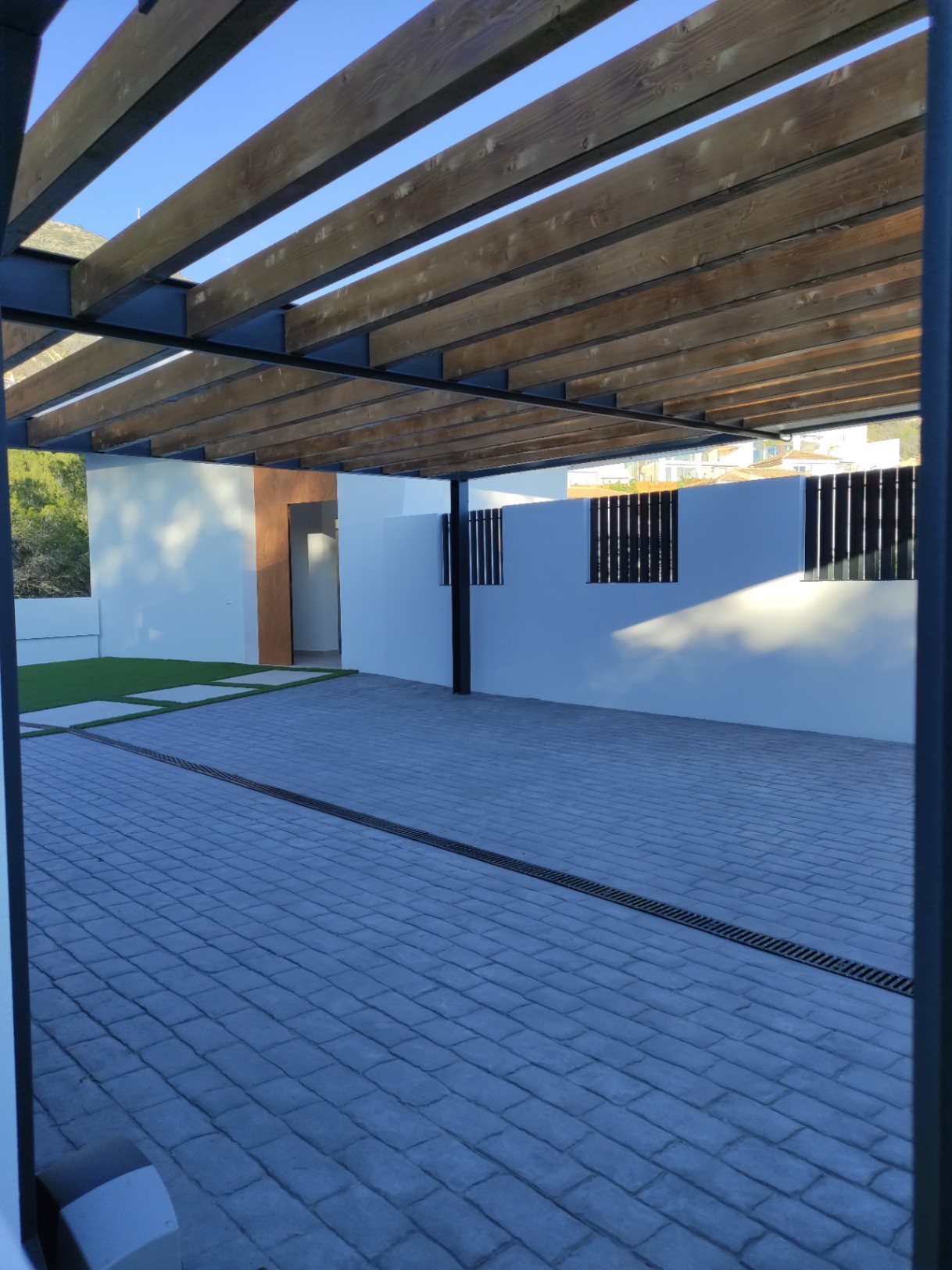 Zona de aparcamiento exterior con pérgola de vigas de madera, suelo de adoquines grises y acceso directo a la vivienda mediante paneles de madera natural.
