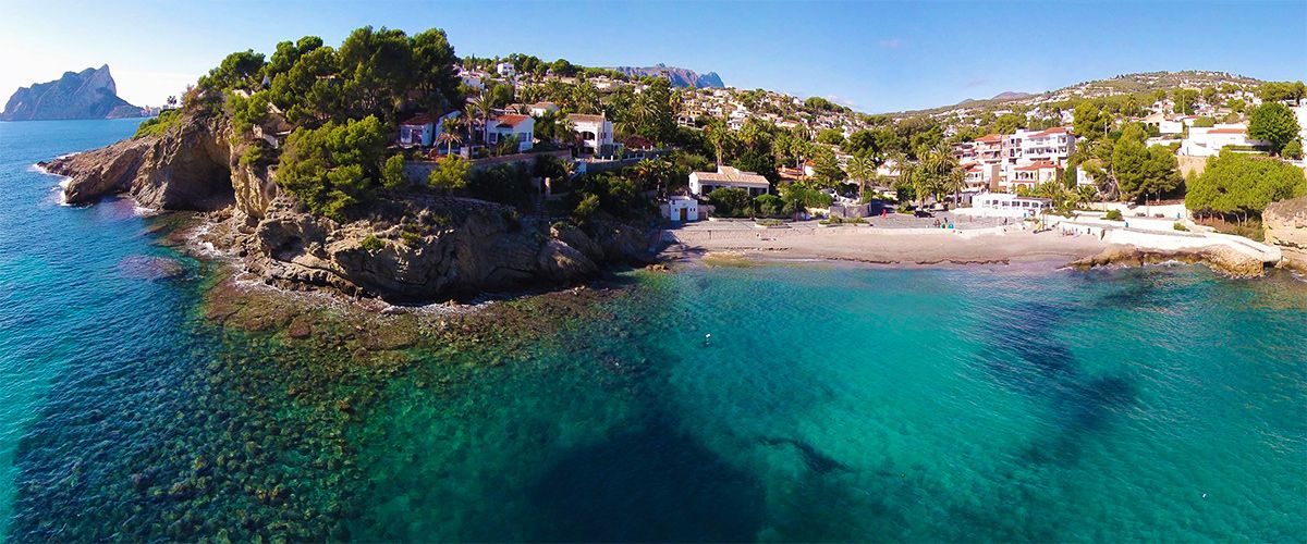 Vista aérea de la playa de La Fustera con aguas turquesas, acantilados rocosos y el Peñón de Ifach al fondo, rodeada de vegetación mediterránea y villas residenciales.