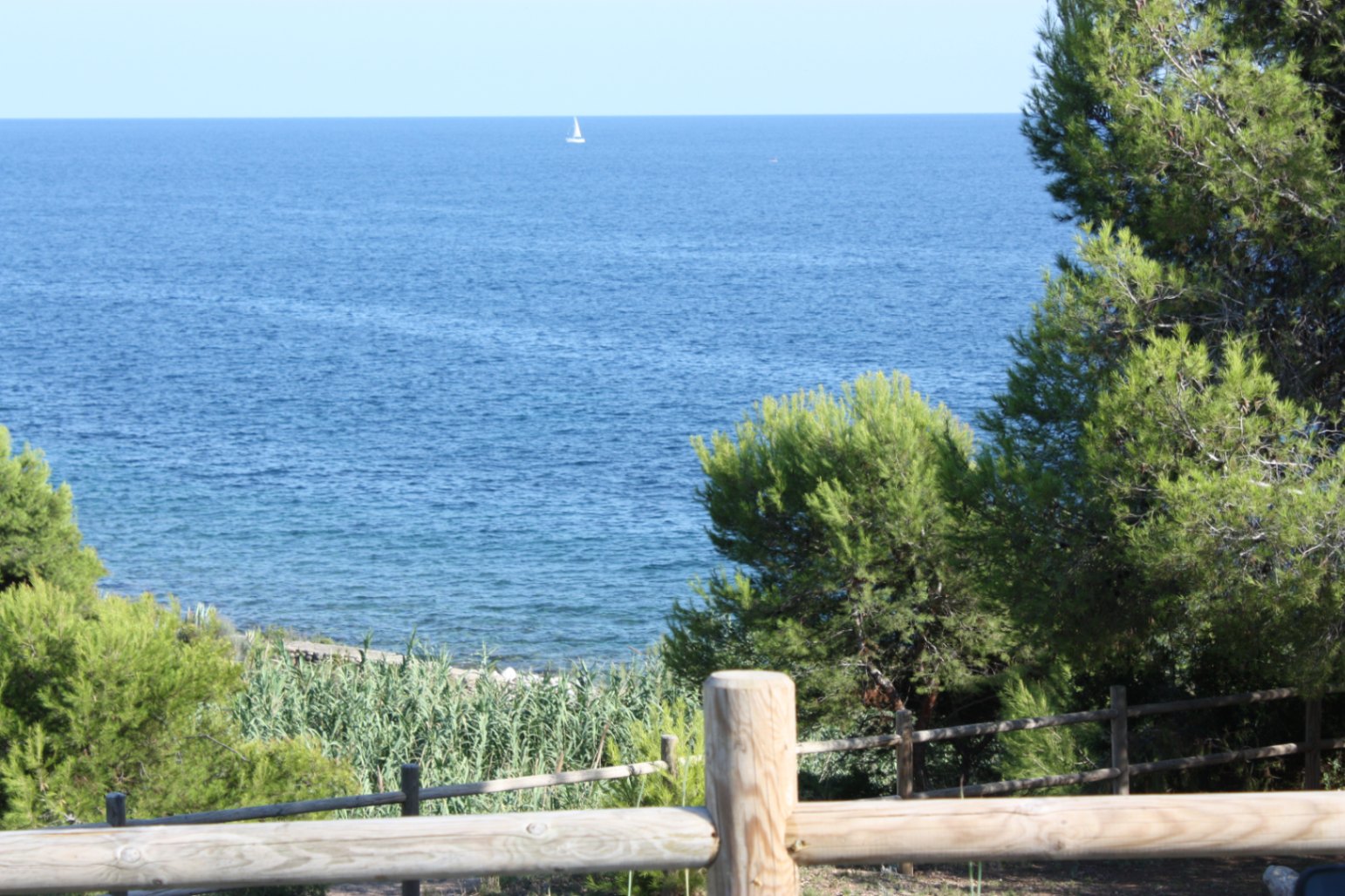 Vista despejada al mar Mediterráneo desde la propiedad, enmarcada por vegetación autóctona y una valla de madera rústica con un velero en el horizonte.