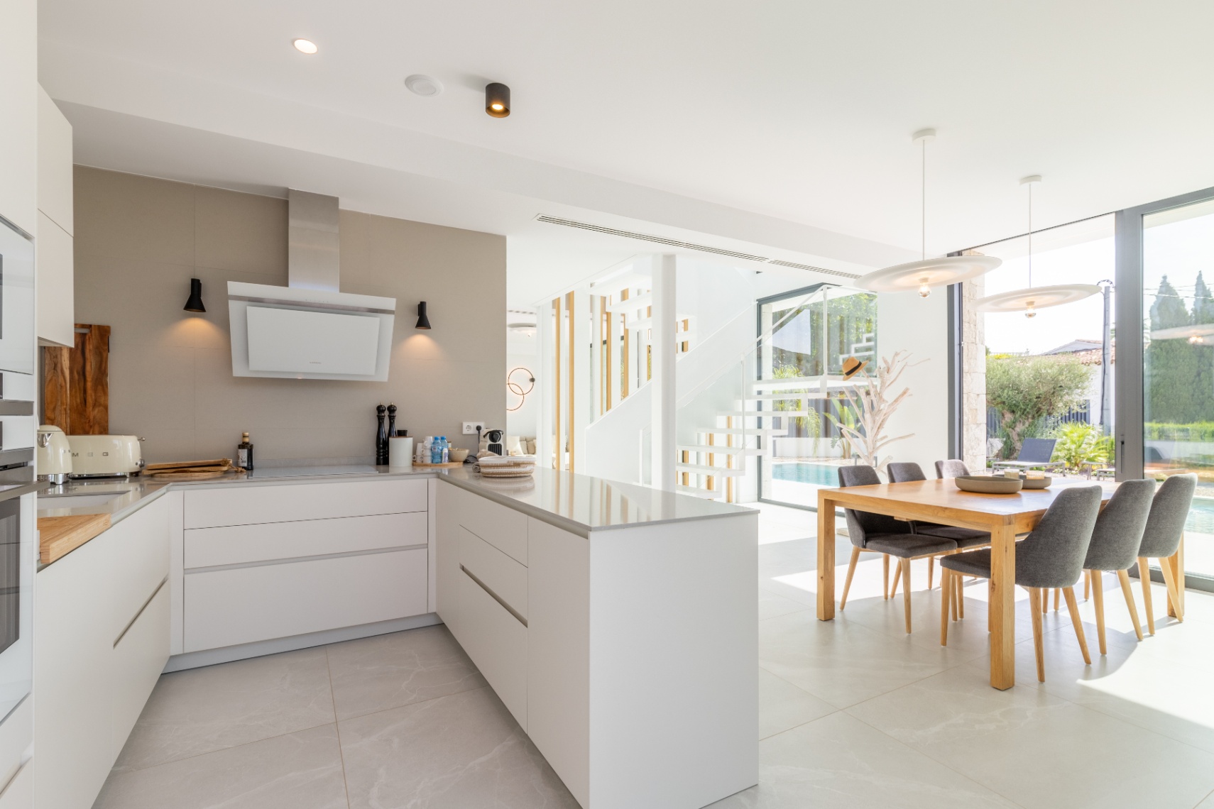 Cocina moderna con isla central, encimeras claras y comedor con vistas a la piscina.