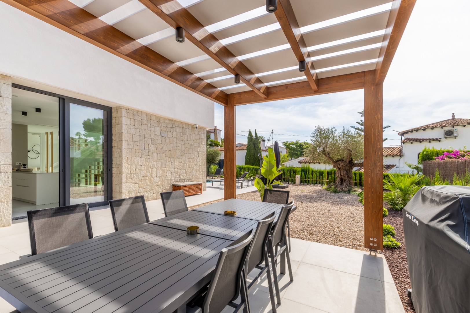 Comedor exterior cubierto con pérgola de madera y vistas al jardín mediterráneo.
