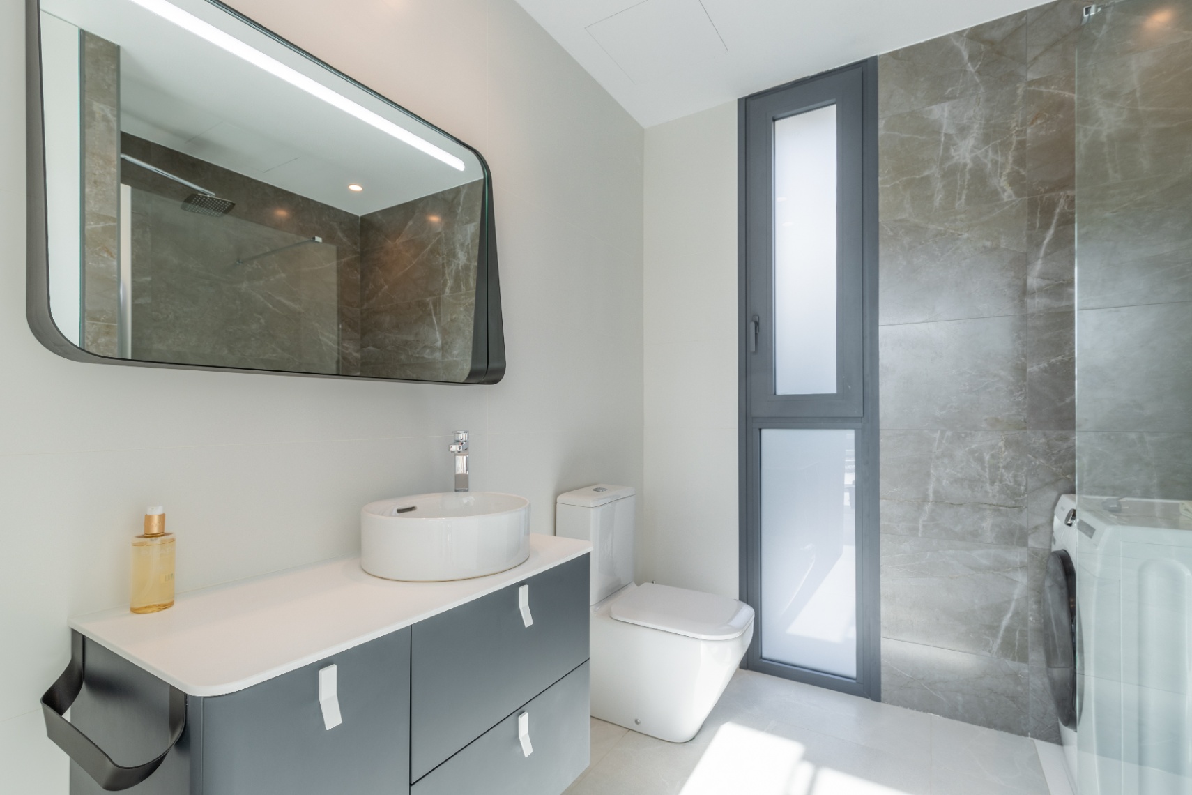Detalle de baño moderno con lavabo cerámico, espejo retroiluminado y revestimiento de mármol gris.