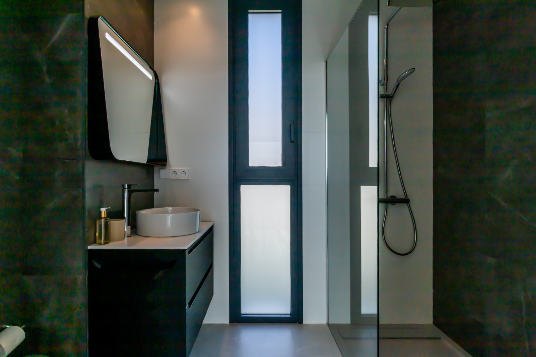 Detalle de baño moderno con lavabo cerámico, mueble negro y ducha con mampara de cristal.
