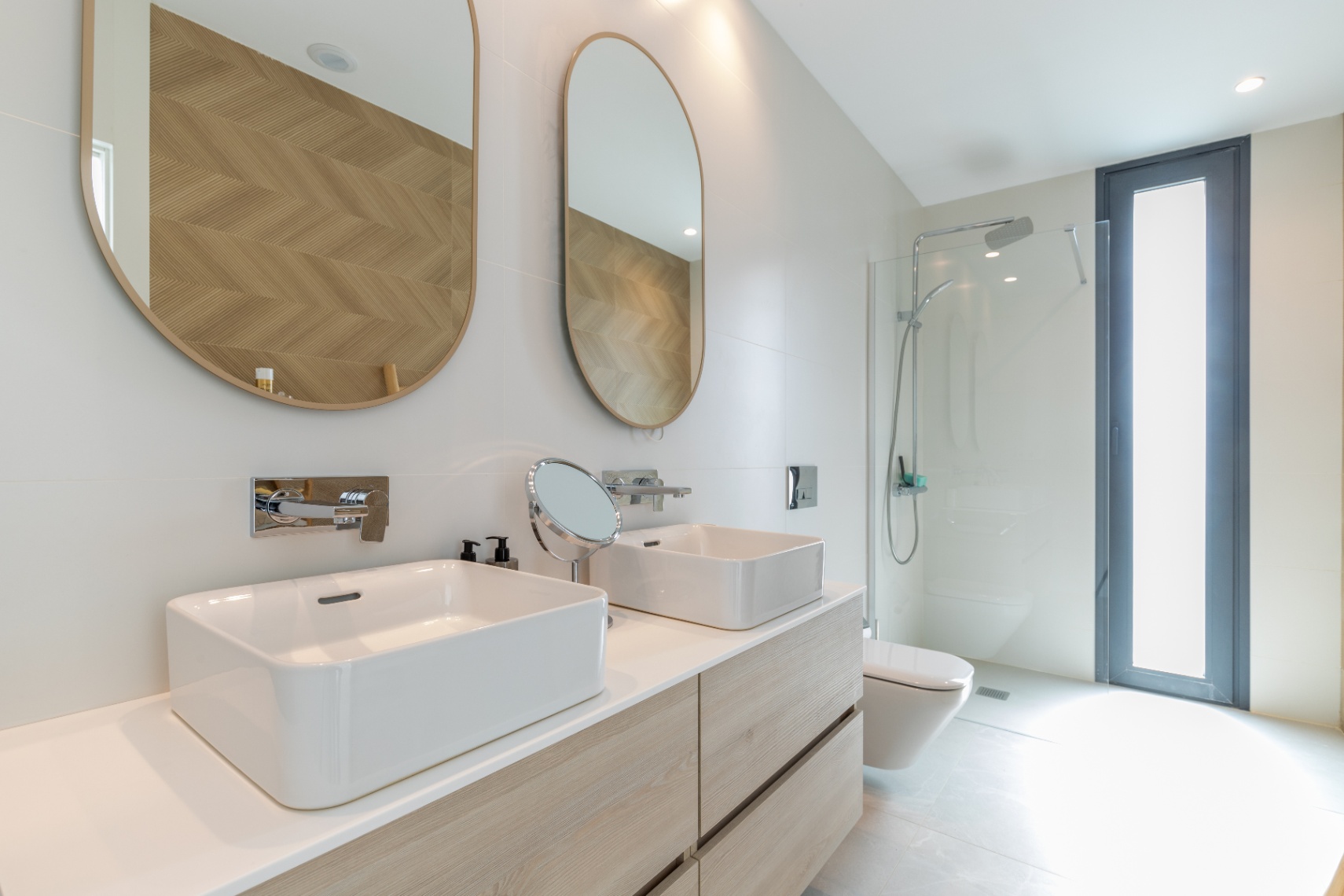 Detalle de baño moderno con lavabo doble, espejos ovalados y ducha con mampara de cristal.