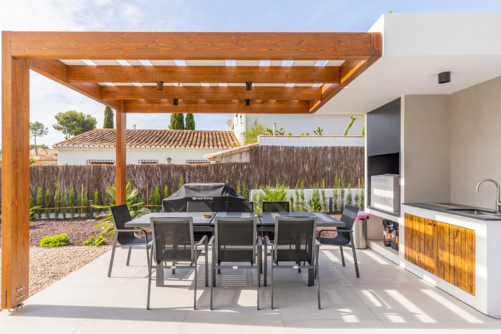 Detalle de la zona de comedor exterior con pérgola de madera y cocina exterior con fregadero y barbacoa.