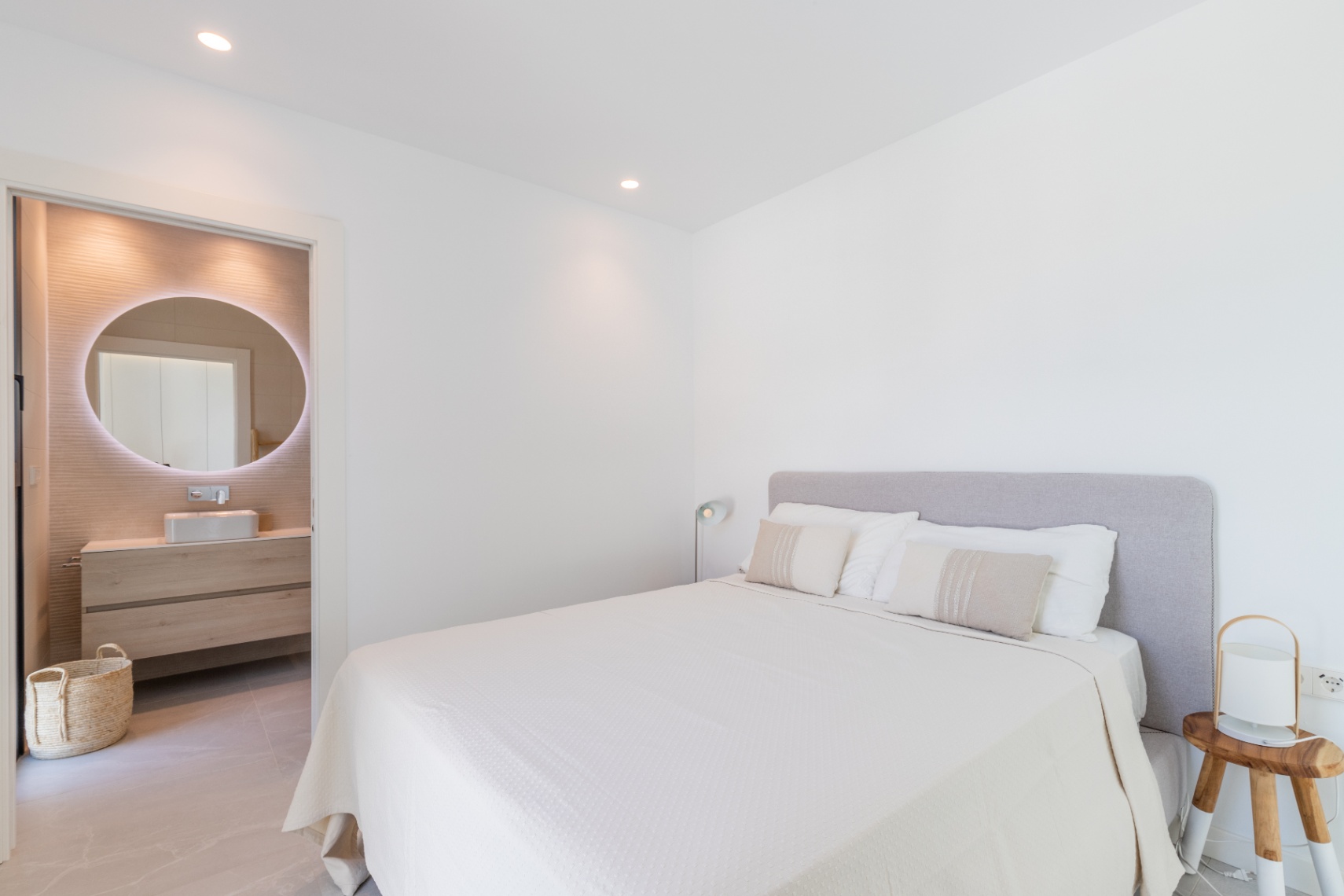Dormitorio luminoso con baño en suite. Cama doble con cabecero gris y ropa de cama blanca. Baño con espejo circular iluminado.