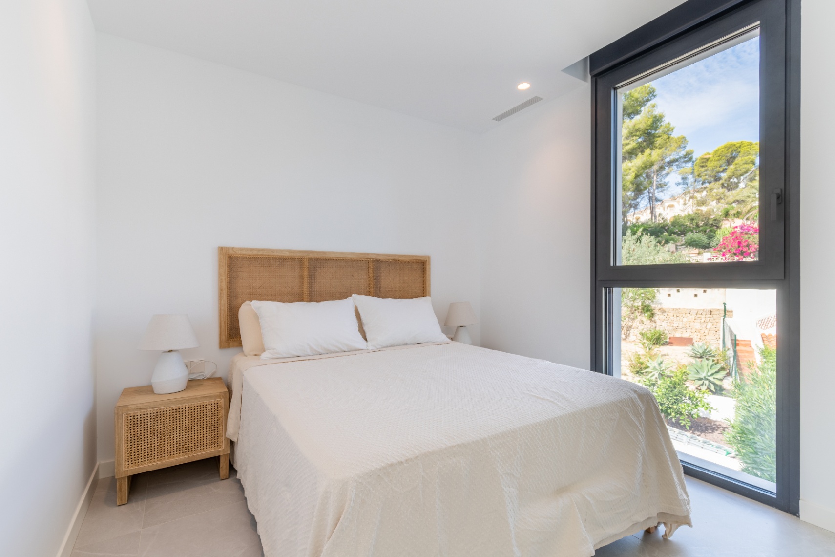 Dormitorio luminoso con cabecero de ratán y vistas al jardín desde la ventana de marco negro.