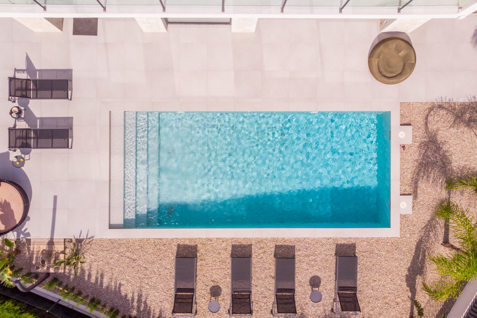 Vista aérea de la piscina rectangular con escalones, rodeada de baldosas claras y grava. Tumbonas y vegetación completan el diseño.