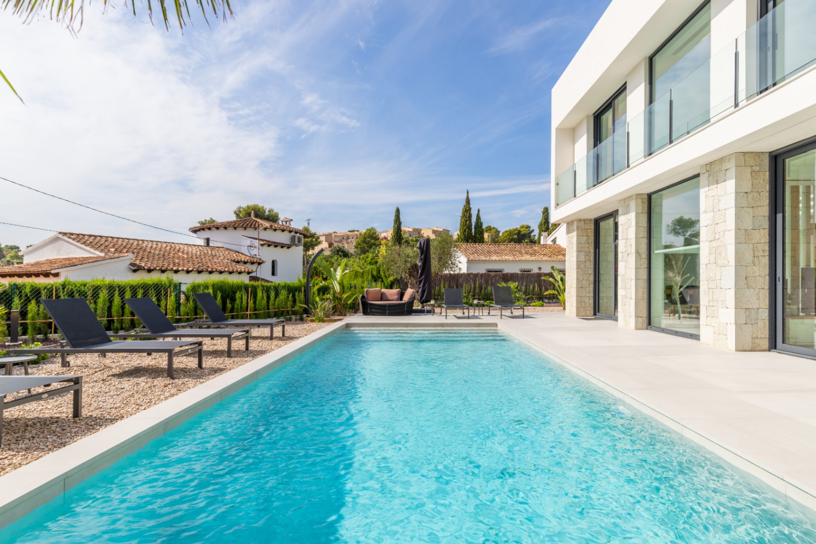 Vista de la piscina turquesa rodeada de tumbonas y la fachada moderna de piedra de la villa.