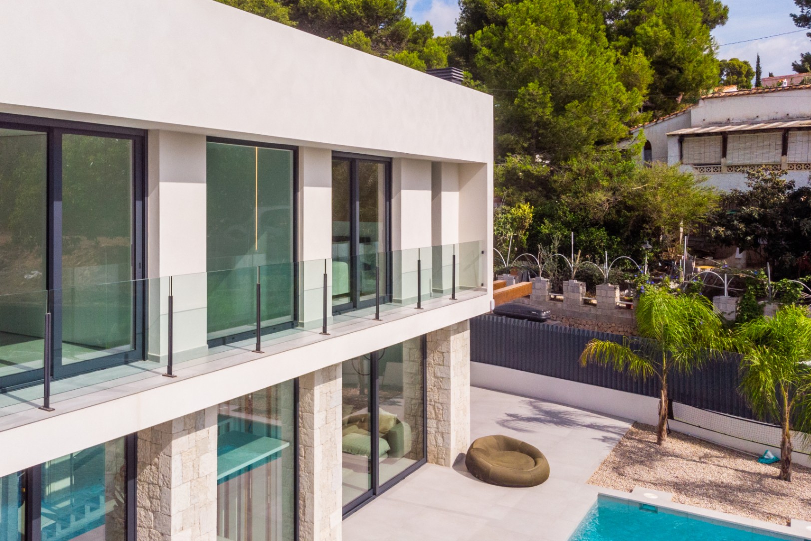 Vista exterior de la villa con balcones de vidrio, revestimiento de piedra y piscina turquesa.