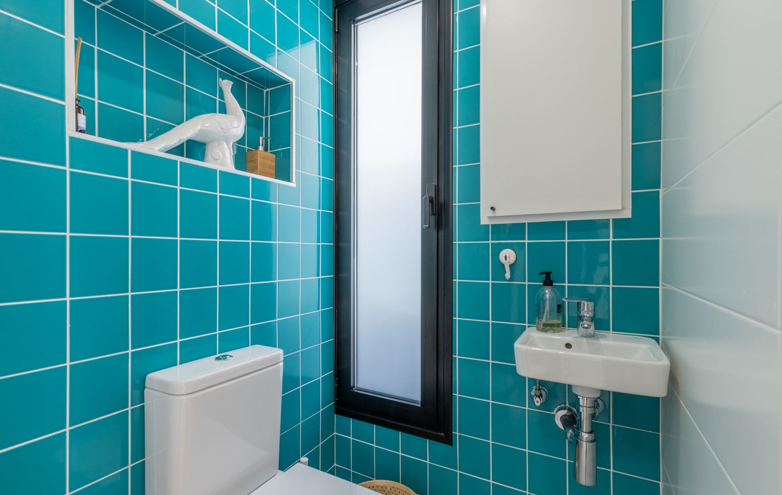 Aseo de cortesía con azulejos turquesa, hornacina decorativa empotrada y lavabo suspendido compacto. Incluye ventana vertical de perfil oscuro y cristal traslúcido.