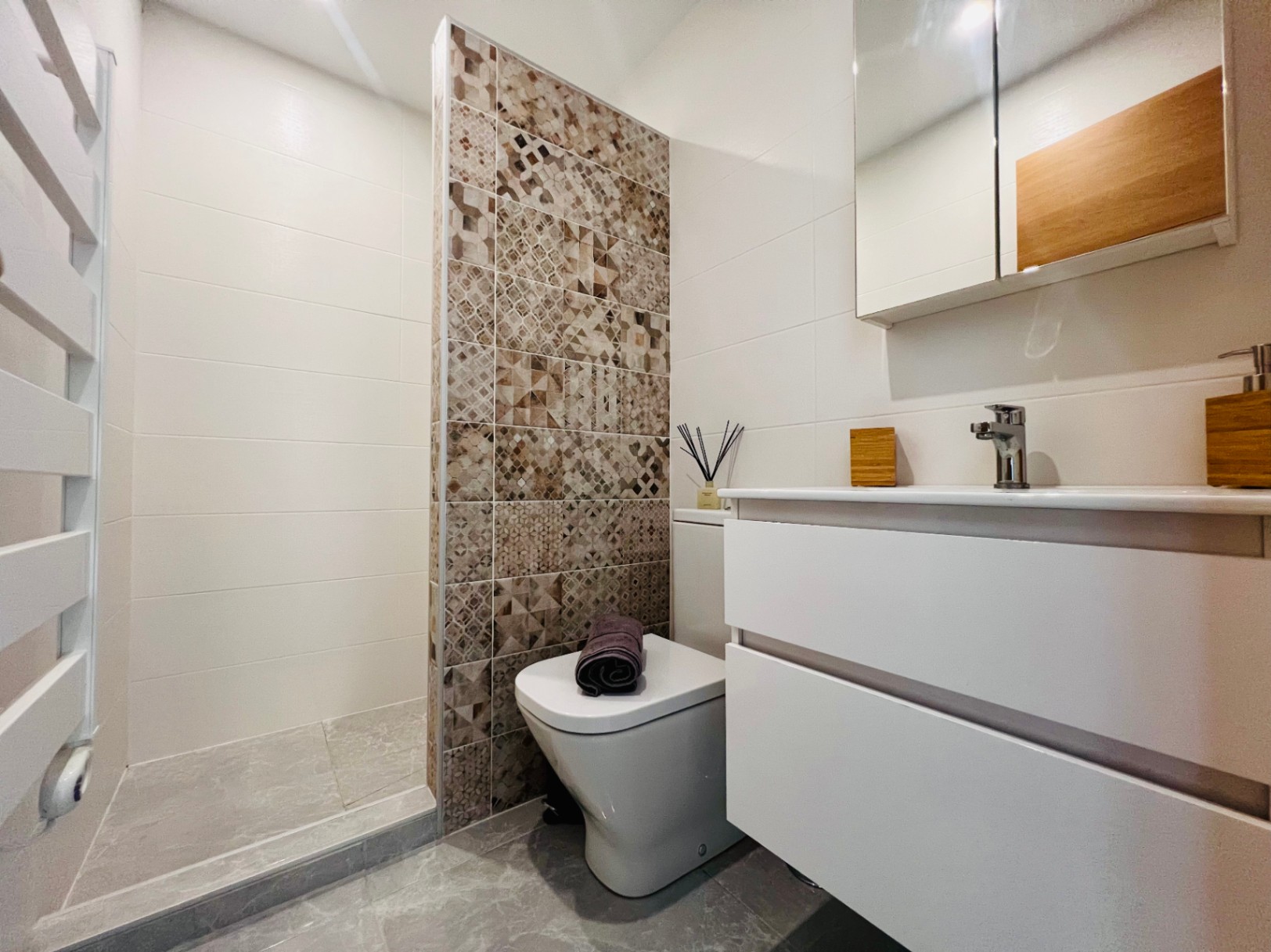 Baño moderno con ducha a ras de suelo, pared decorativa de azulejos geométricos, mueble de lavabo blanco suspendido y radiador toallero integrado.