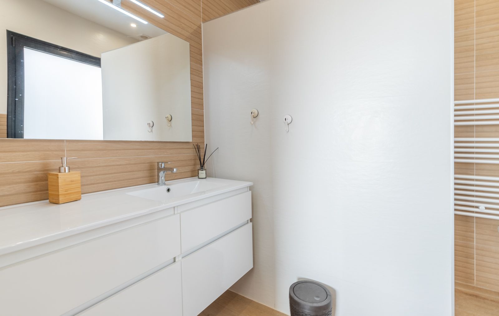 Baño moderno con mueble suspendido blanco, encimera con lavabo integrado y revestimiento de pared con efecto madera. Incluye espejo con iluminación LED y radiador toallero.