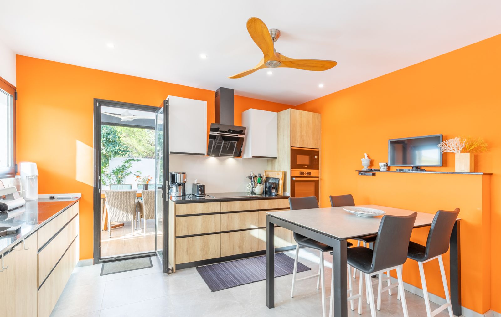 Cocina moderna con paredes naranja vibrante, mobiliario de madera clara y zona de comedor integrada con acceso directo a la terraza exterior.