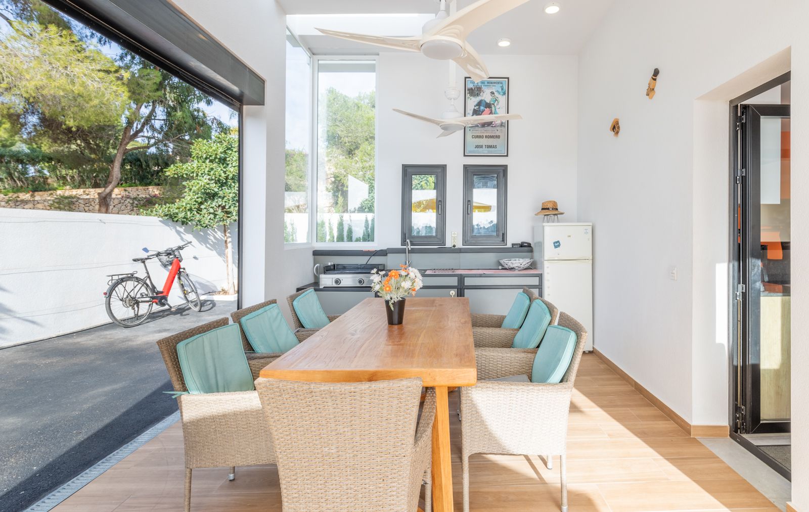 Comedor exterior cubierto con cocina de verano, mesa de madera maciza, sillas de mimbre y ventiladores de techo, integrado con el patio mediante grandes ventanales.