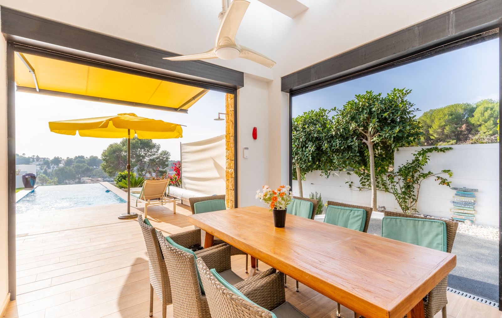 Comedor exterior cubierto con mesa de madera y sillas de mimbre, situado junto a la piscina infinita y equipado con ventilador de techo y toldos automáticos.