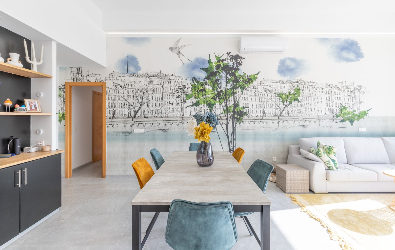 Comedor moderno con mesa de efecto piedra, sillas de terciopelo en azul y mostaza, y un mural artístico de ciudad. Suelos de gres gris y acceso al salón.