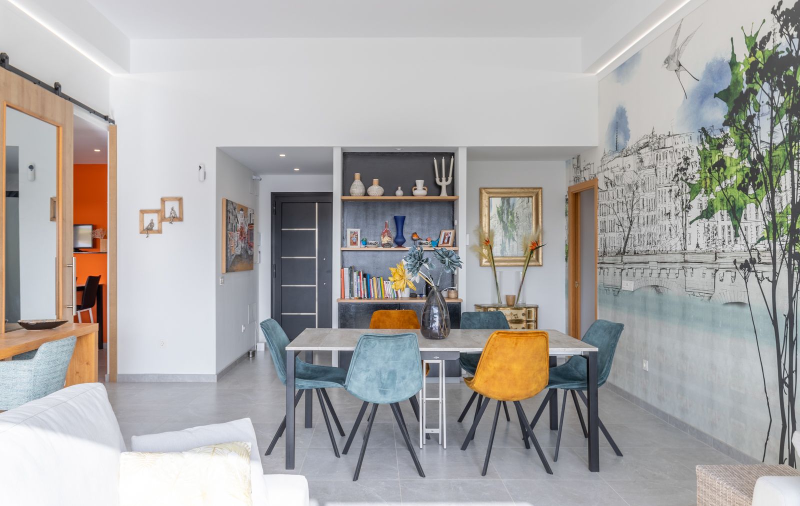 Comedor moderno con mesa minimalista y sillas de terciopelo en tonos azul y ocre. Destaca una pared artística con mural ilustrado y estantería integrada bajo iluminación LED.