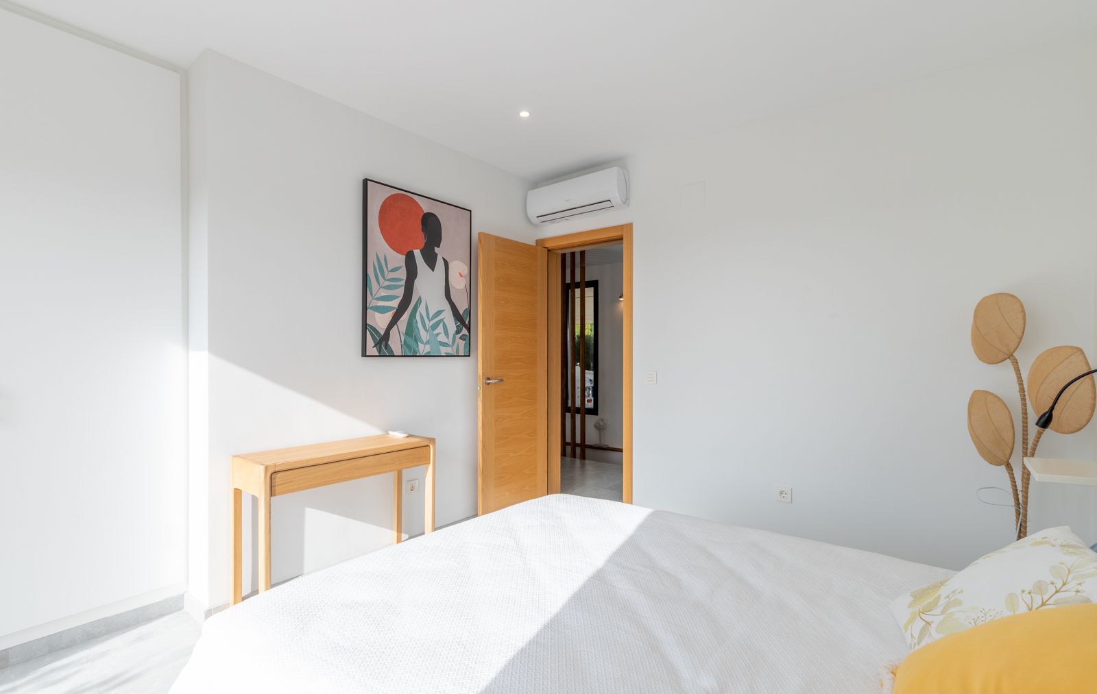 Dormitorio moderno con paredes blancas, escritorio de madera natural y aire acondicionado. Incluye carpintería de madera clara y acceso directo al pasillo.