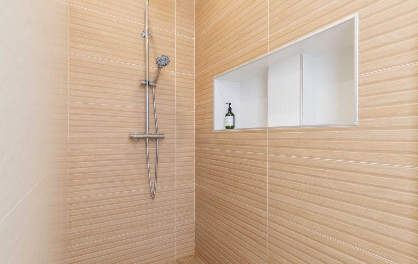 Ducha de diseño con revestimiento cerámico de efecto madera horizontal, grifería cromada y un práctico nicho de pared integrado con acabado blanco.
