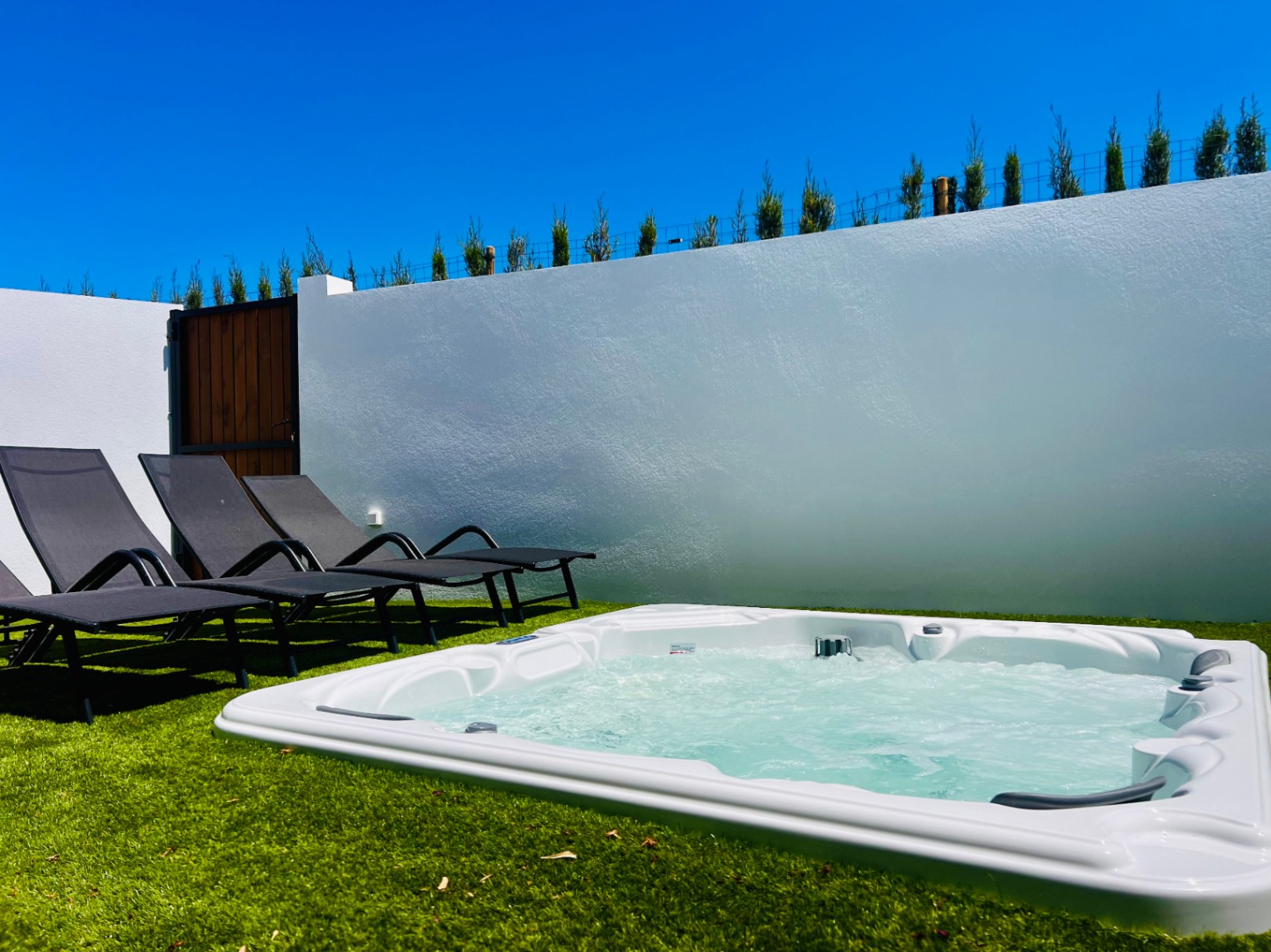 Jacuzzi privado integrado en césped artificial con tumbonas de diseño, rodeado por un muro blanco que garantiza total privacidad bajo el cielo despejado.
