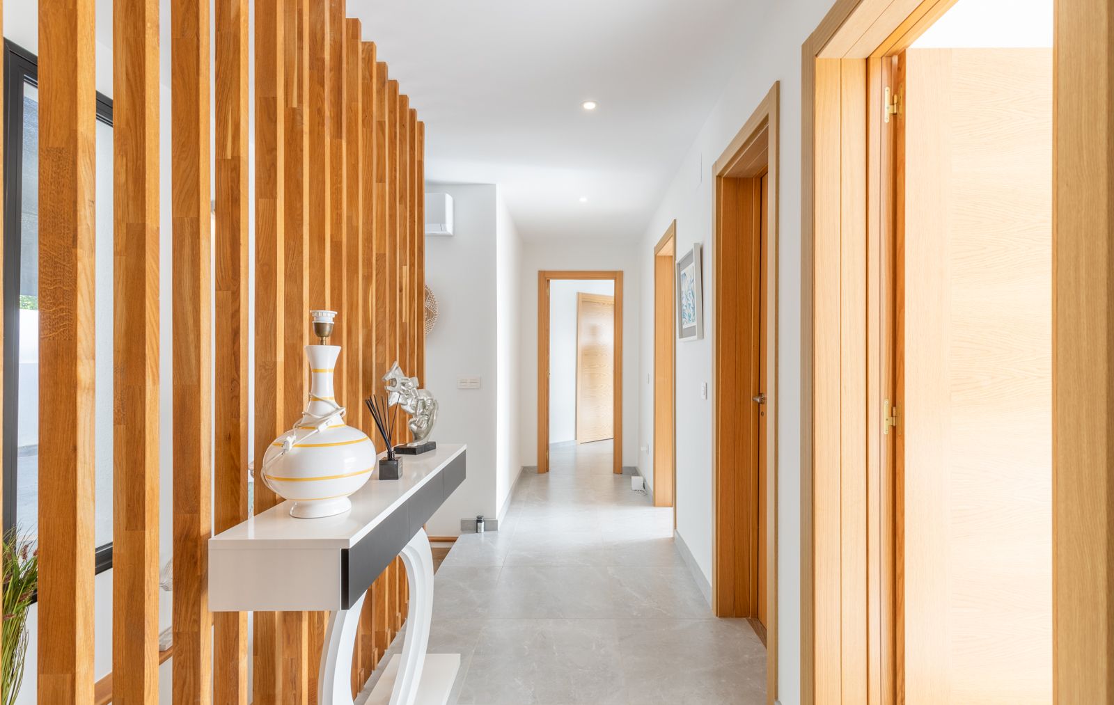 Pasillo moderno con separador de lamas de madera natural, suelos de gres porcelánico gris y carpintería interior en madera clara coordinada.
