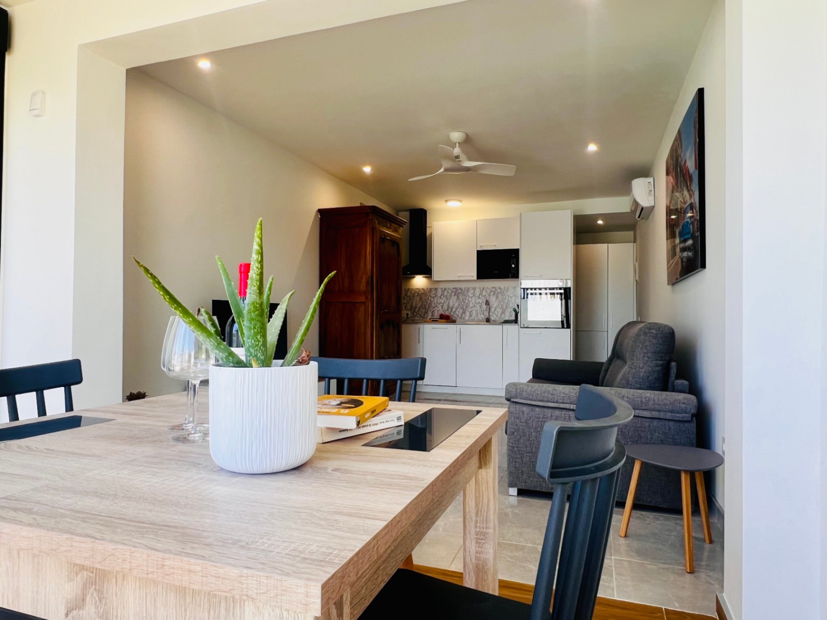 Salón-comedor integrado con cocina blanca moderna, sofá gris, ventilador de techo y un armario de madera clásica que aporta contraste al diseño contemporáneo.