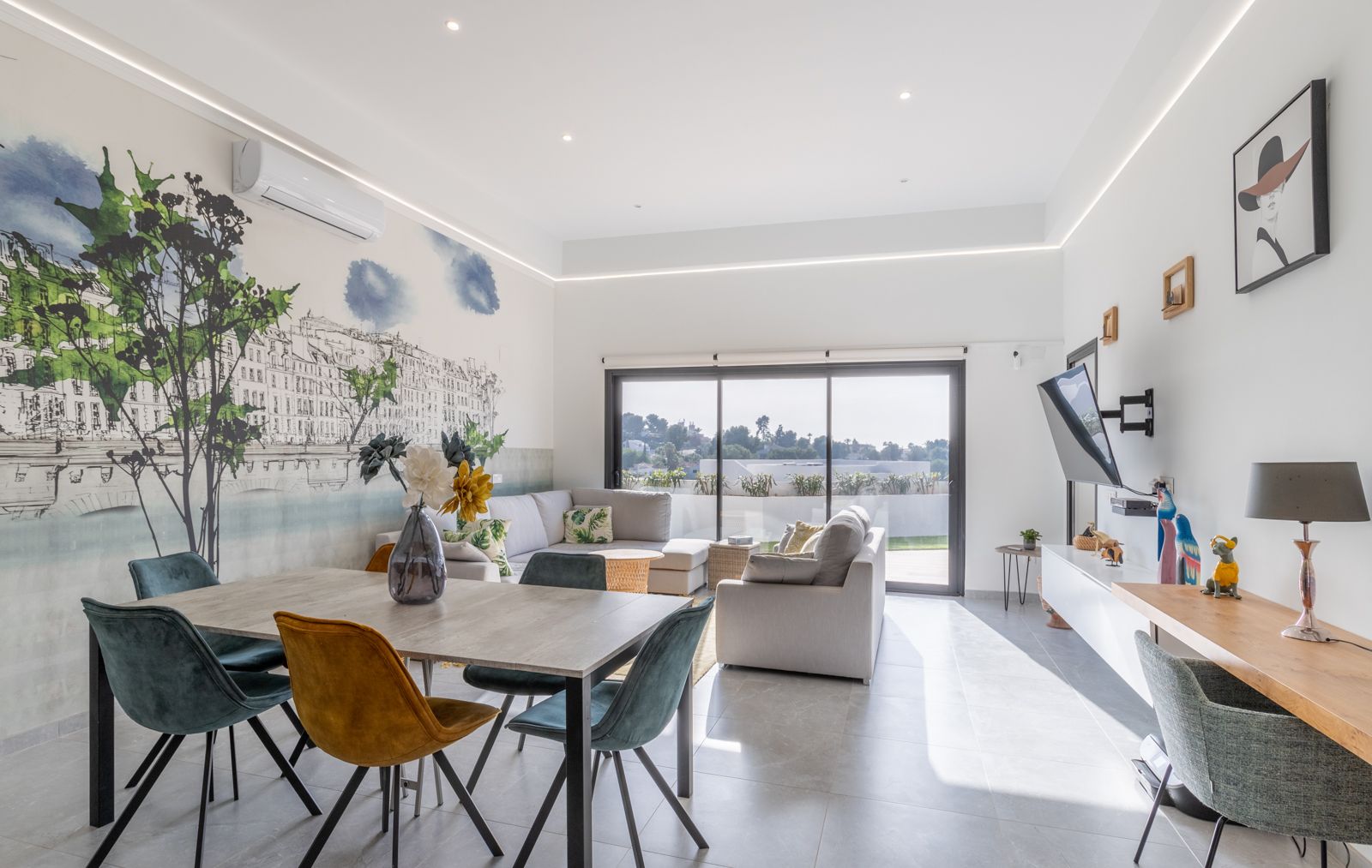 Salón-comedor moderno con mural artístico, mobiliario contemporáneo en tonos mostaza y azul, y acceso directo a la terraza mediante amplios ventanales.