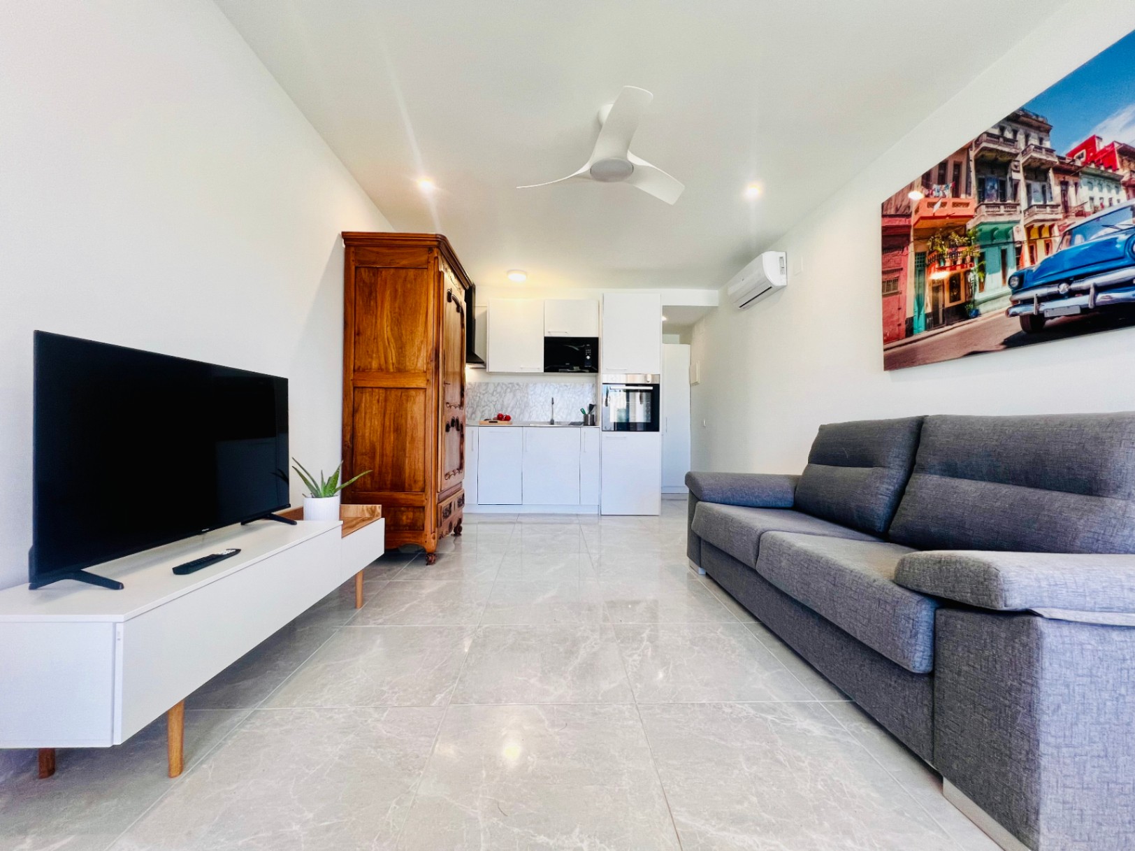 Salón de uno de los apartamentos privados con suelos de gres porcelánico, sofá gris, cocina integrada blanca y un armario de madera clásica que aporta contraste.