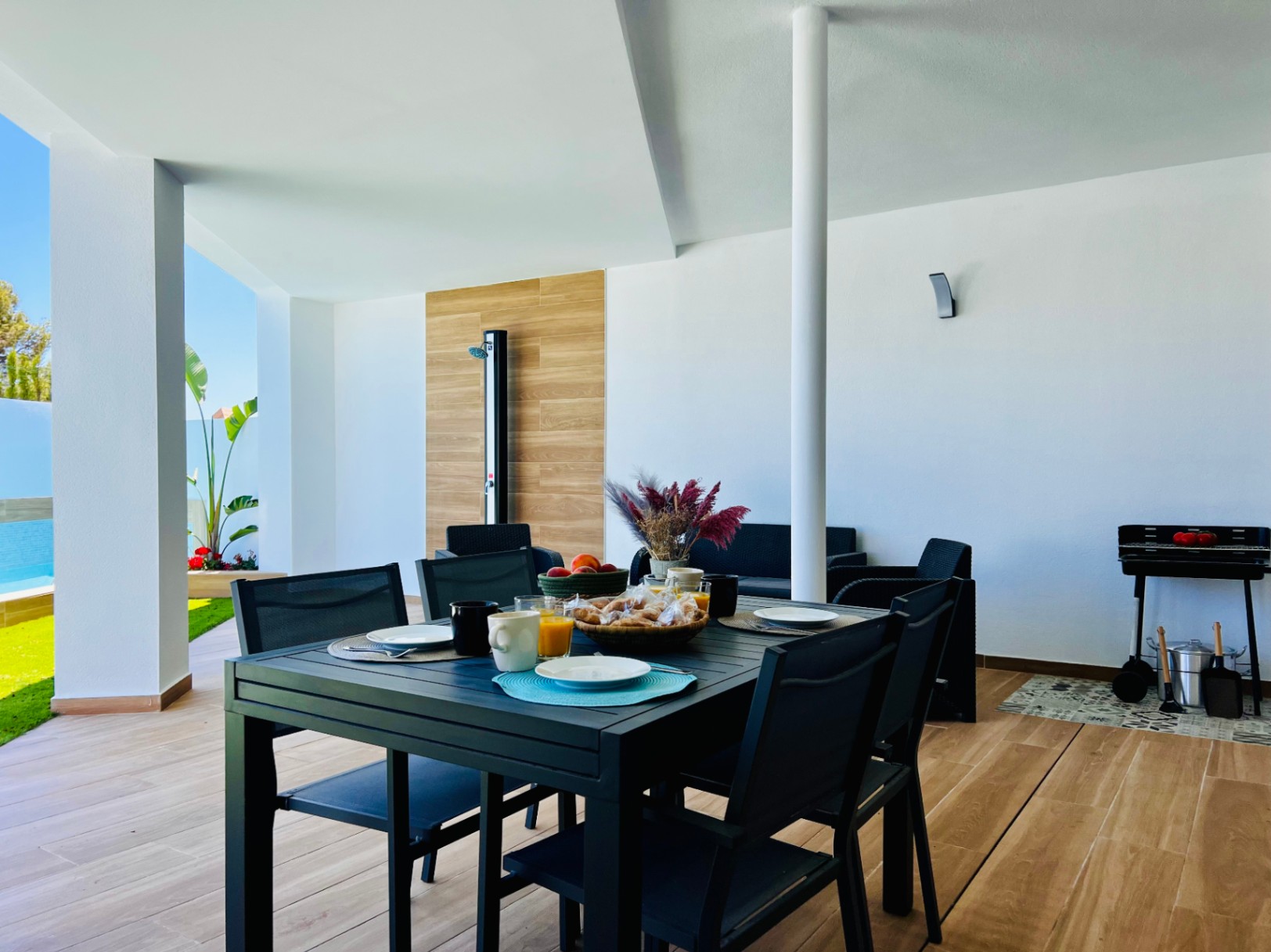 Terraza cubierta con suelos de gres porcelánico efecto madera, zona de comedor exterior, barbacoa y ducha exterior integrada en panel decorativo.