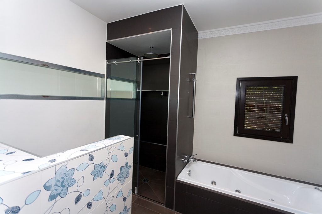 Baño contemporáneo con bañera integrada y cabina de ducha independiente. Presenta azulejos en contraste chocolate y crema, con un murete decorativo de motivos florales.