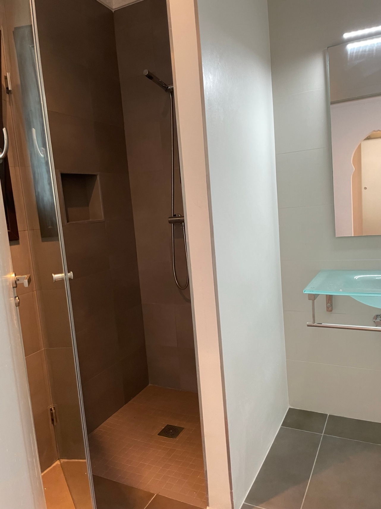 Baño moderno con ducha de obra revestida en tonos tierra, hornacina integrada y lavabo de cristal suspendido con toallero cromado.