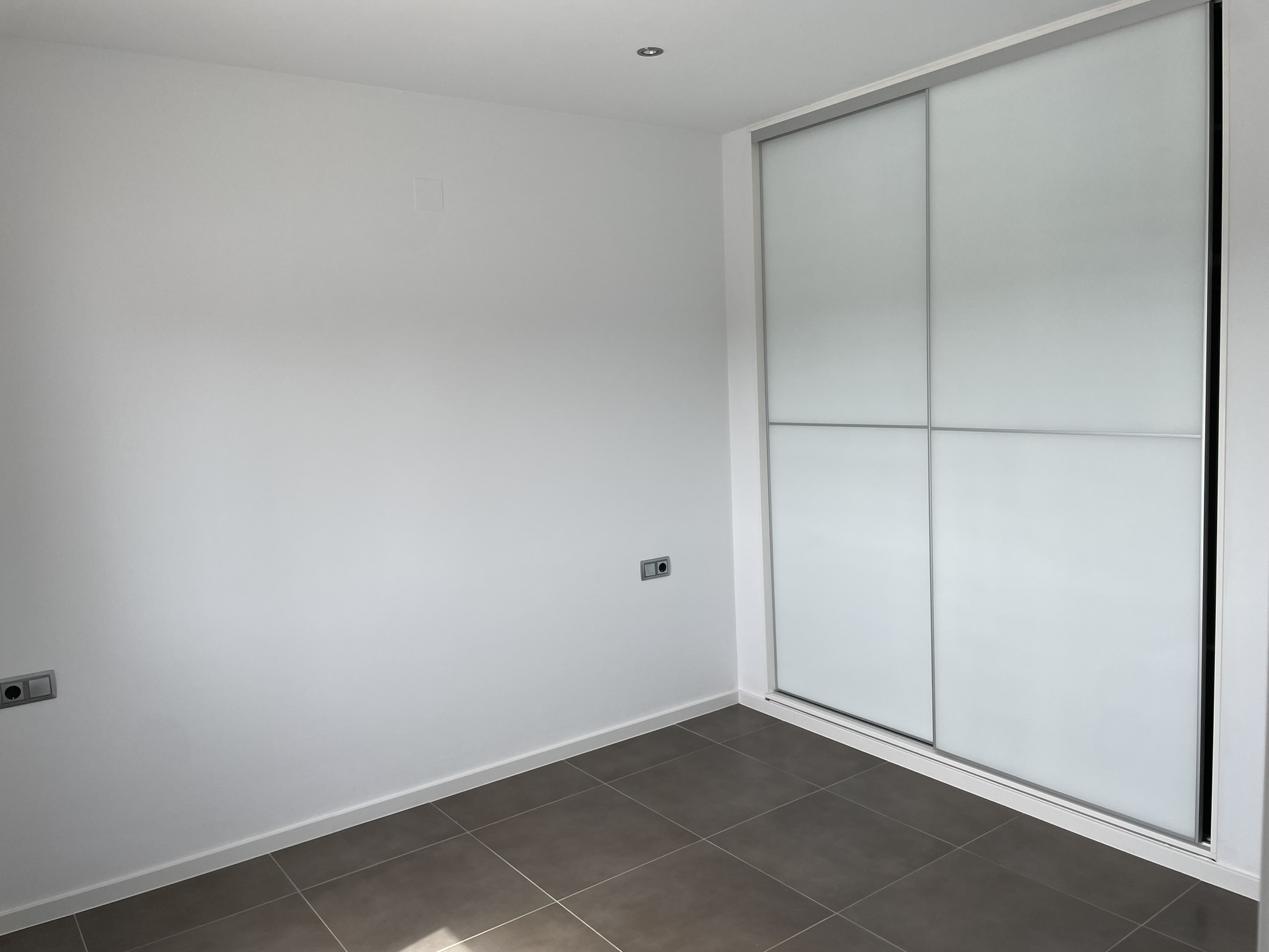Dormitorio con armario empotrado de puertas correderas blancas, suelos de gres porcelánico oscuro y focos encastrados en el techo.