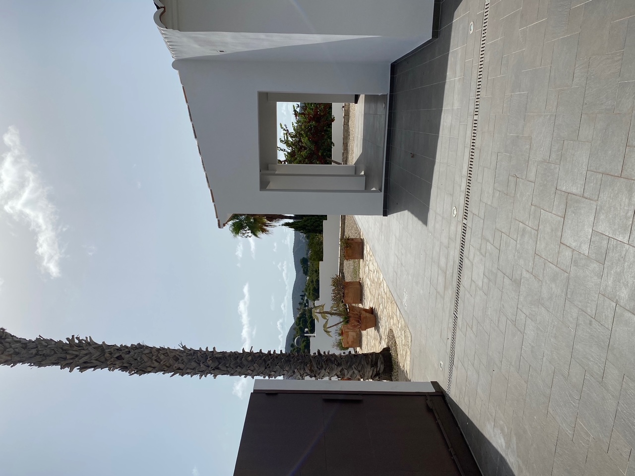 Entrada exterior con pavimento gris, puerta metálica, palmera y porche cubierto. Incluye iluminación empotrada en el suelo y vistas despejadas a las montañas.