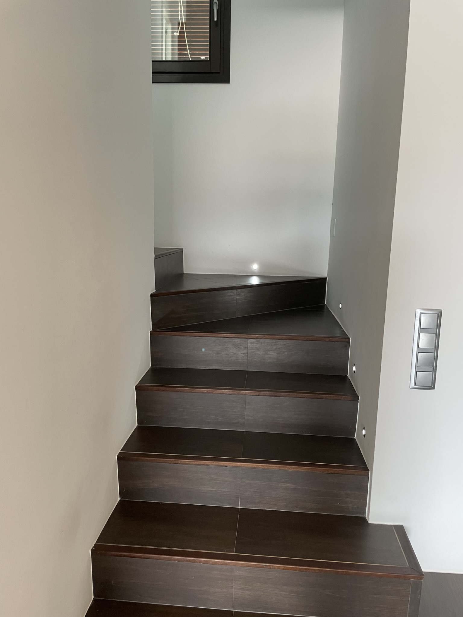 Escalera interior de gres porcelánico oscuro con iluminación LED integrada en la pared y paredes blancas lisas que conducen a la planta superior.