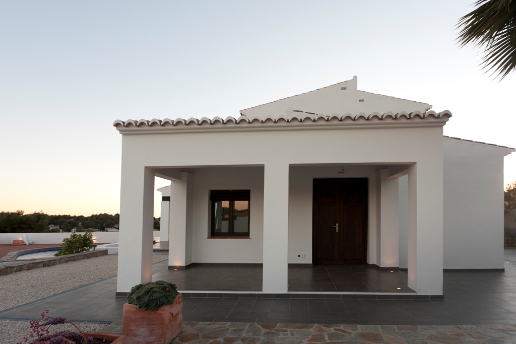 Fachada blanca con porche cubierto, puerta de madera antigua restaurada e iluminación integrada en el suelo, junto a zona de grava y piscina.
