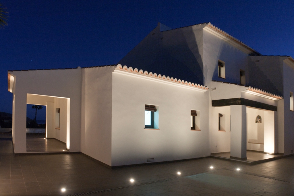 Fachada blanca de estilo mediterráneo iluminada al anochecer con focos empotrados en el suelo. Destacan las tejas curvas y el porche con vigas oscuras sobre pavimentos cerámicos.