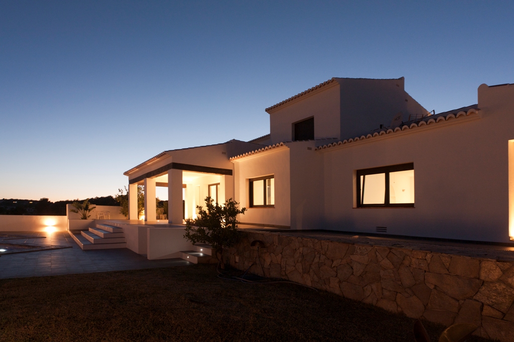 Fachada exterior de la villa al atardecer, destacando la iluminación cálida, los muros de piedra natural y las amplias terrazas que conectan con la zona de piscina.