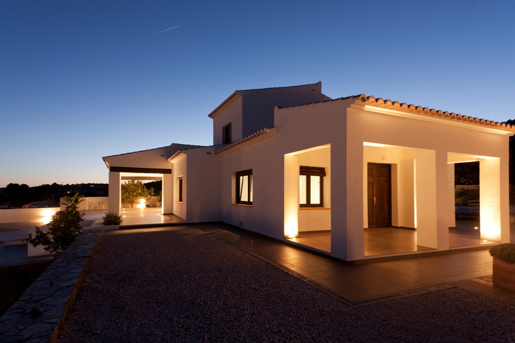 Fachada mediterránea al atardecer con iluminación integrada en pilares, destacando la puerta de madera restaurada y los acabados en blanco bajo un cielo crepuscular.
